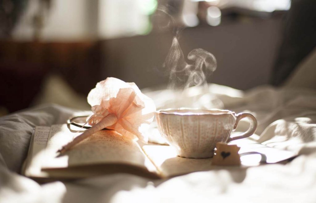 Ilustračný obrázok: čaj - hrnček s čajom, z ktorého stúpa horúca para položený na knihe na posteli s ozdobným umelým kvetom
