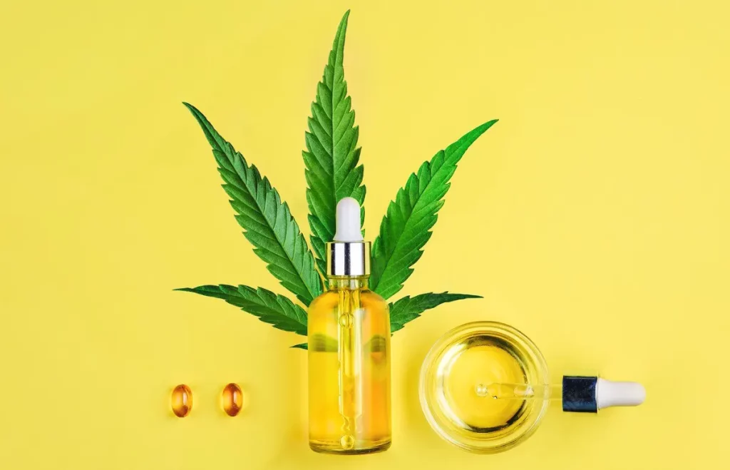 CBD produkty na žltom pozadí ozdobené konopným listom: flakón s CBD olejom, kapsule a nádobka s pipetou